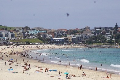 A wide-angle, high-elevation view of a crowded public beach on a sunny day. Hundreds of people are scattered across the light-colored sand and swimming in the turquoise ocean water. The background features a densely packed hillside of multi-story coastal apartment buildings and houses under a hazy sky.